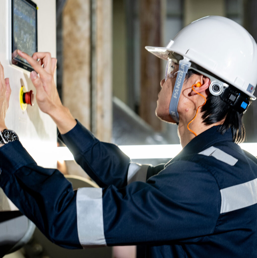 A person wearing a white hard hat, safety goggles, and ear protection operates a touchscreen control panel in an industrial setting.
