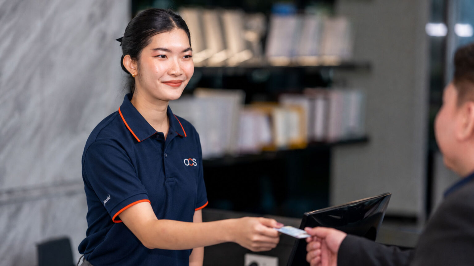A woman in a navy polo shirt smiles as she hands a card to a person across a reception desk, with shelves of books in the background.
