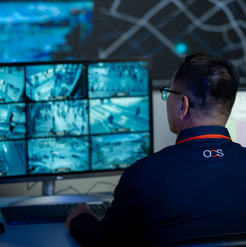 A man in an OCS uniform monitors multiple surveillance camera feeds on two computer screens in a control room.