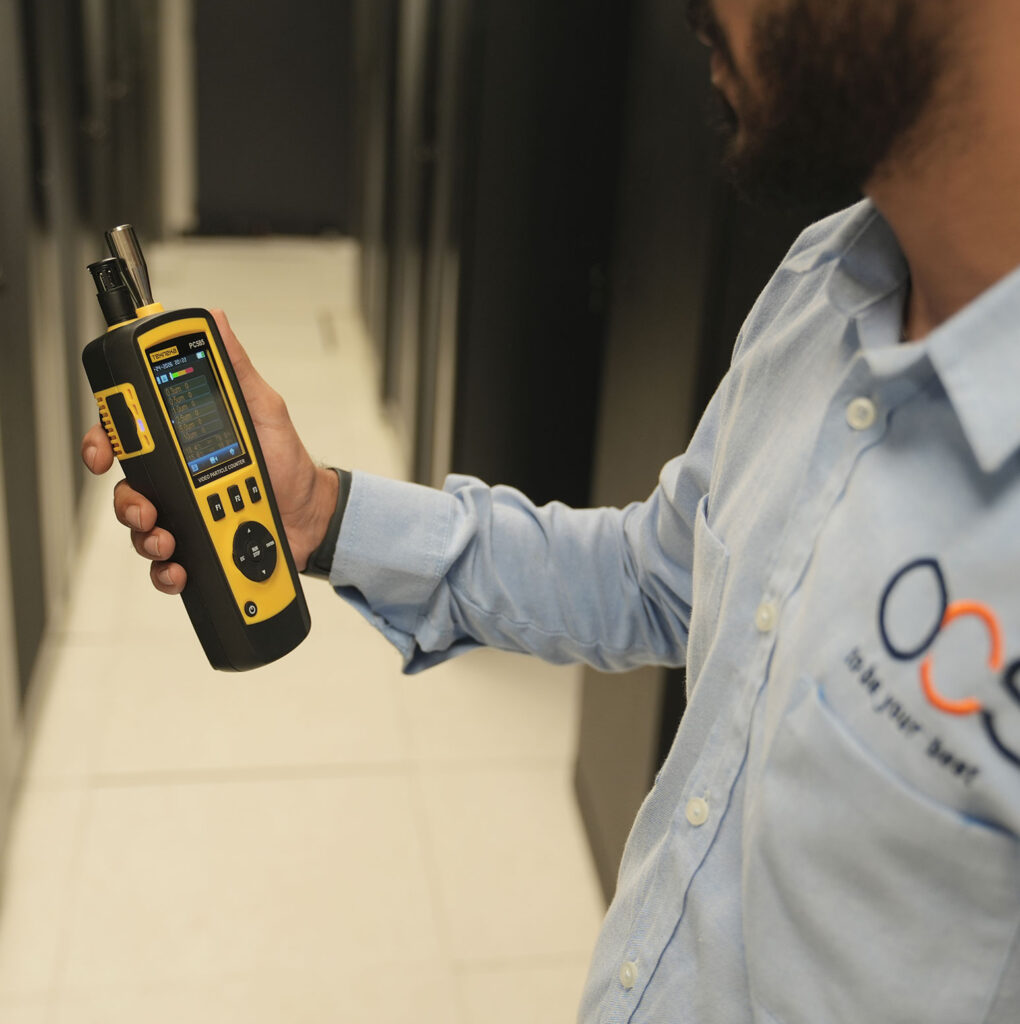A person in a light blue shirt holds a yellow and black handheld device, possibly for measuring air quality, in a room with black cabinets and a white floor.