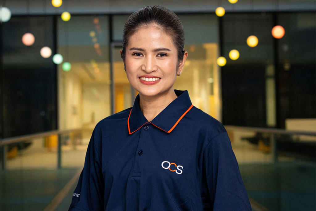 A woman in a navy blue OCS uniform shirt smiles at the camera. She stands indoors with blurred colorful lights and glass walls in the background.