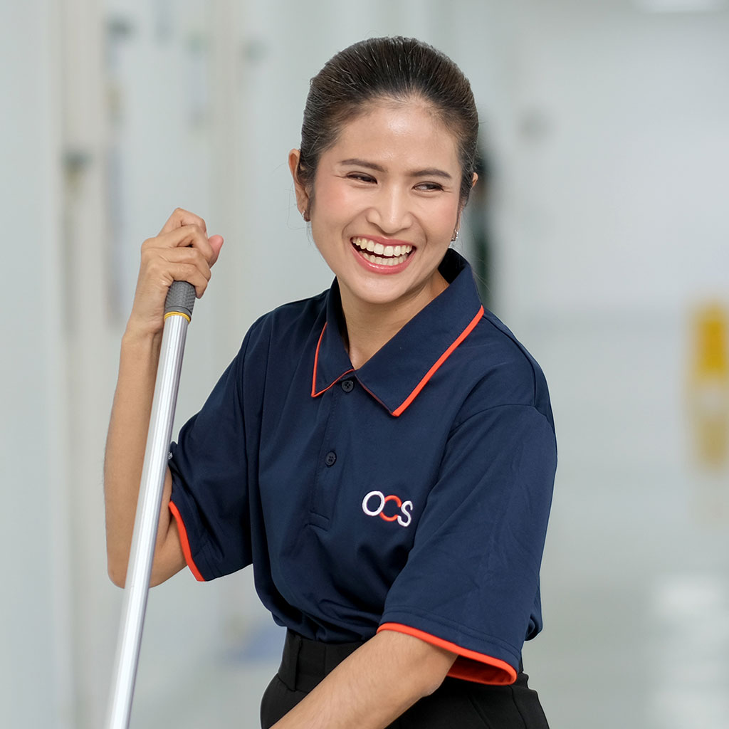 A smiling woman in a navy blue OCS uniform holds a mop in a bright hallway, appearing happy and engaged in her work.