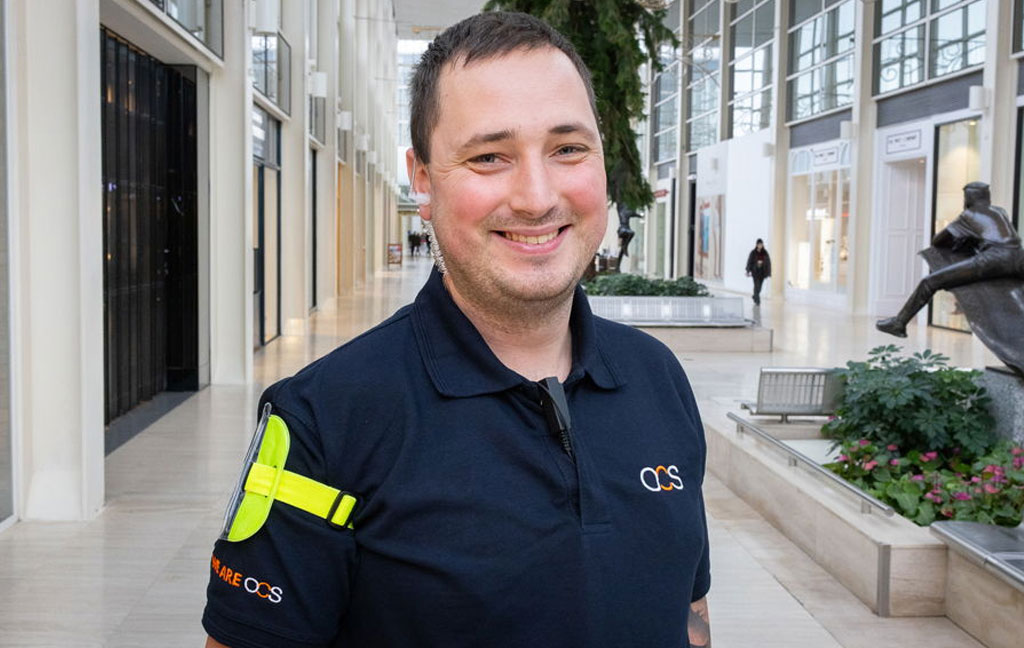 A smiling security guard wearing a dark blue OCS uniform stands inside a modern shopping mall with tall glass walls and indoor plants.
