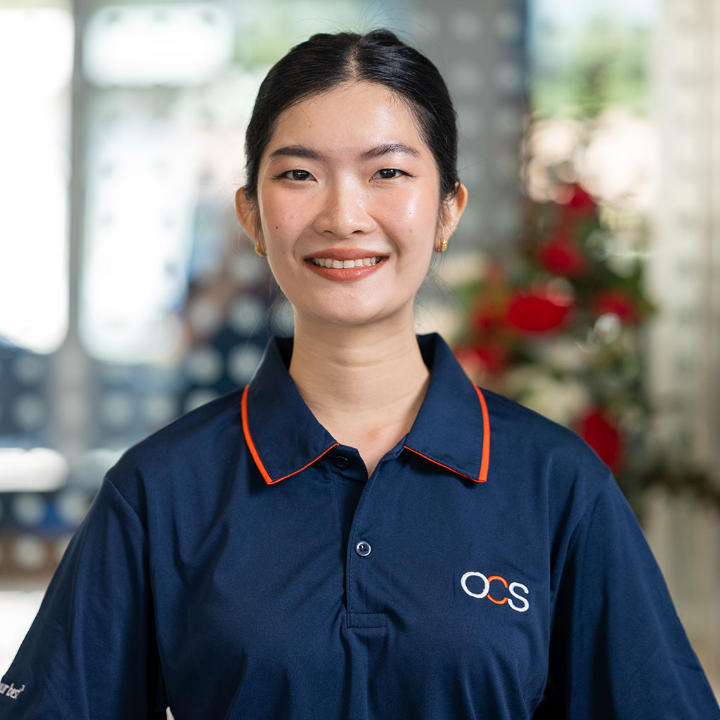 A woman with dark hair tied back smiles at the camera, wearing a navy blue collared shirt with an OCS logo. The blurred background features flowers and bright light.