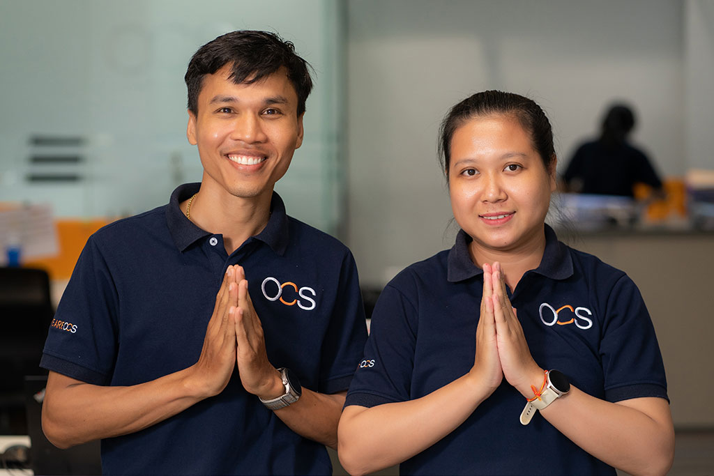 Two people wearing matching navy blue OCS shirts stand side by side indoors, smiling and holding their hands together in a traditional greeting gesture.