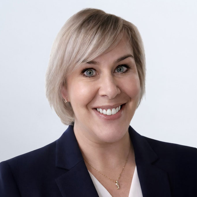 Smiling woman with short blonde hair wearing a dark blazer, white blouse, and a gold necklace, posed in front of a plain light background.