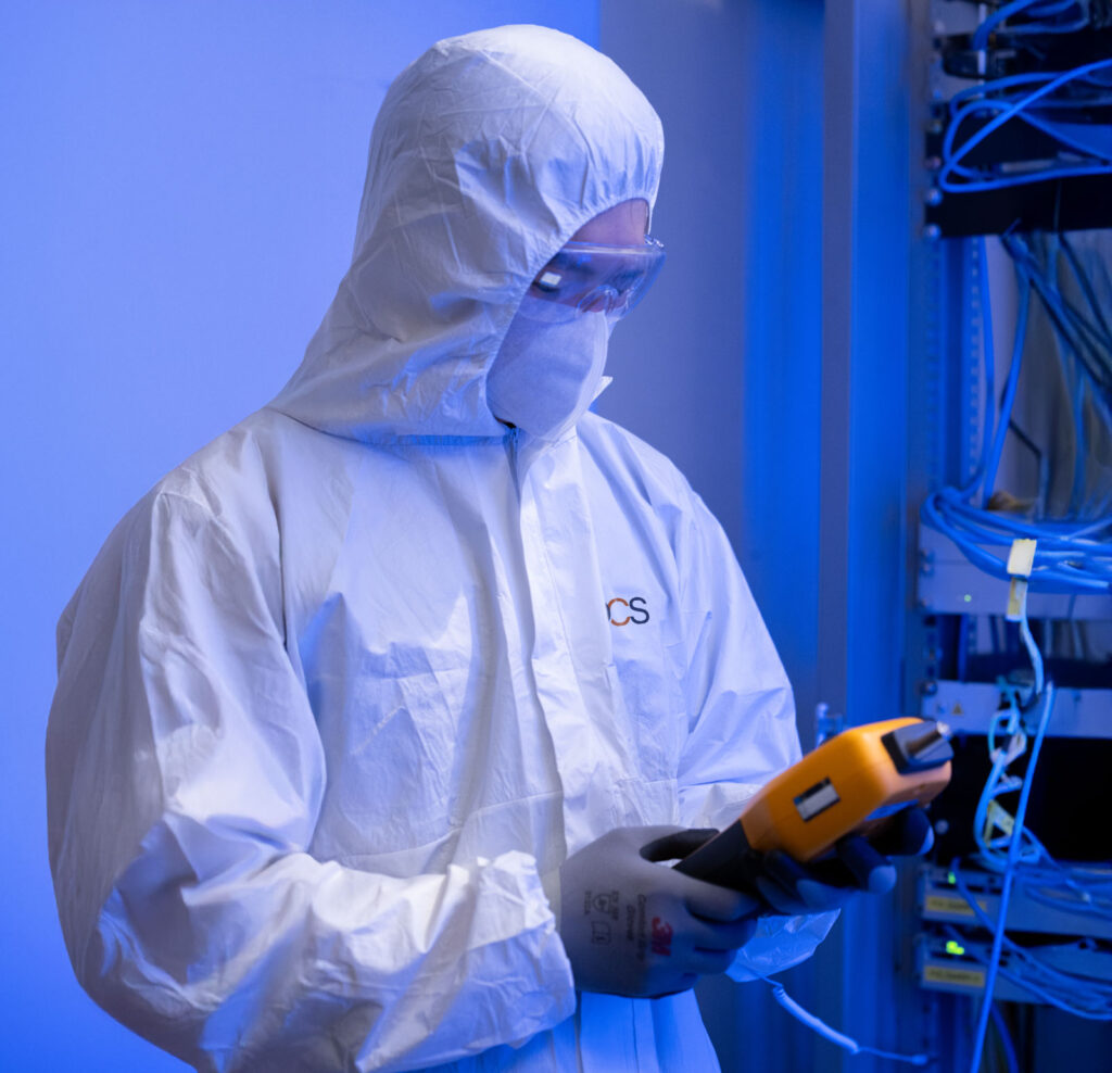 A person in a white protective suit and gloves inspects a yellow electronic device, standing near blue network cables in a lab or tech environment.