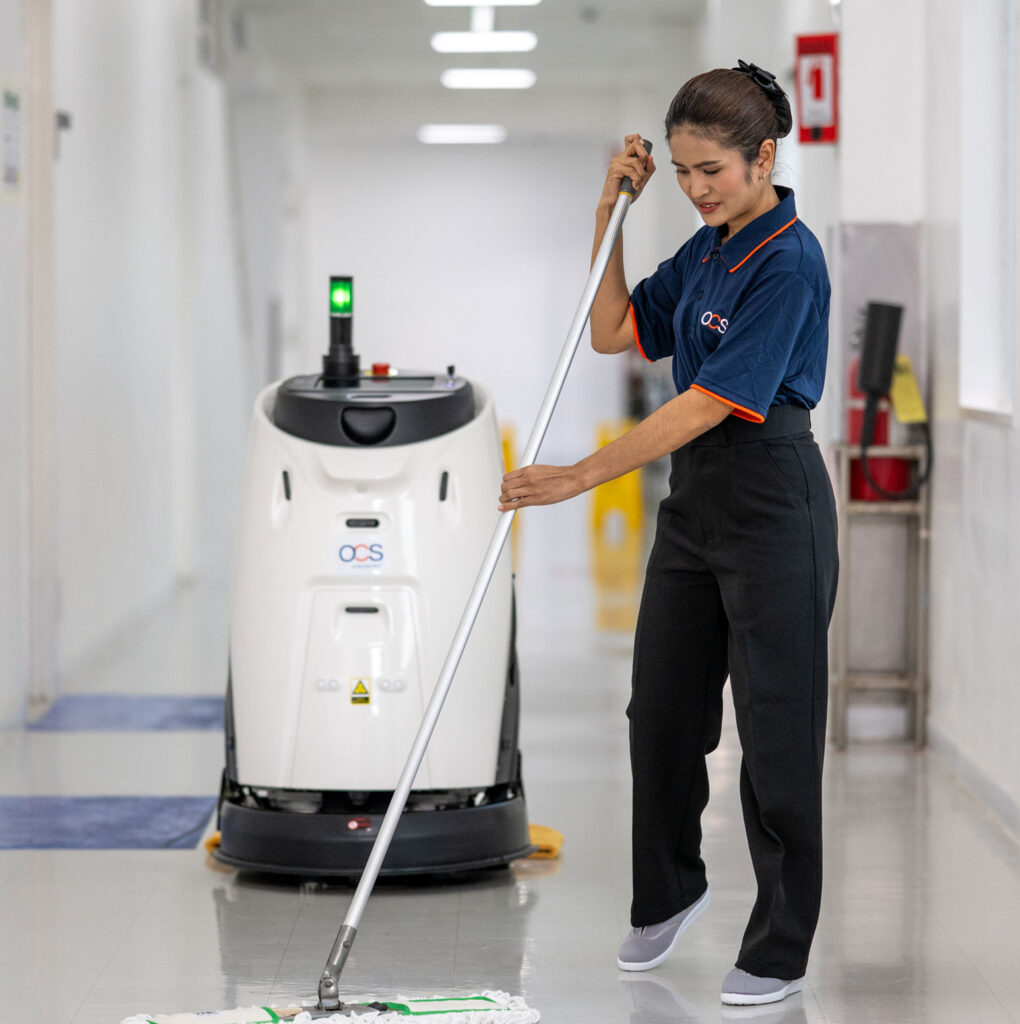 A woman in uniform mops a hallway floor while a large cleaning robot is positioned behind her. The corridor is well-lit and clean, with safety signs and cleaning equipment visible in the background.