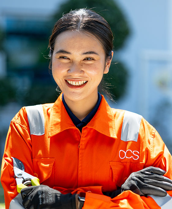 A woman in an orange OCS uniform smiles with arms crossed, holding pruning shears. She is outdoors with greenery and sunlight in the background.
