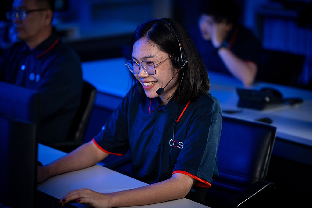 A woman wearing glasses and a headset smiles while working at a computer in an office with dim blue lighting. Two colleagues are visible in the background.