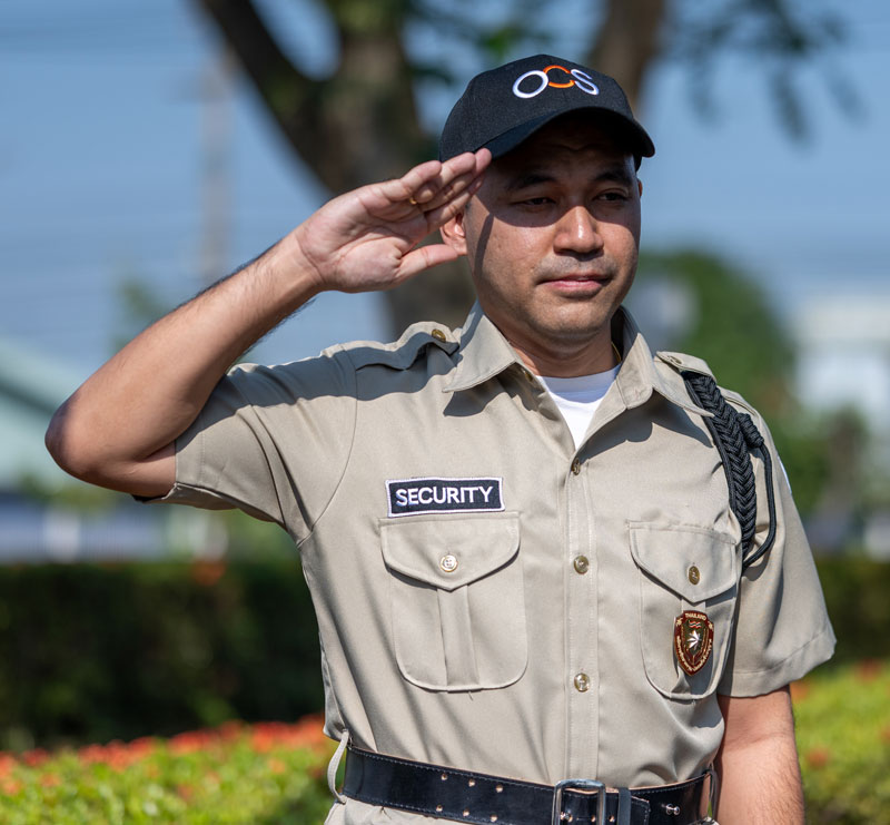 A security guard in a khaki uniform and black cap salutes outdoors on a sunny day, with trees and greenery in the blurred background.