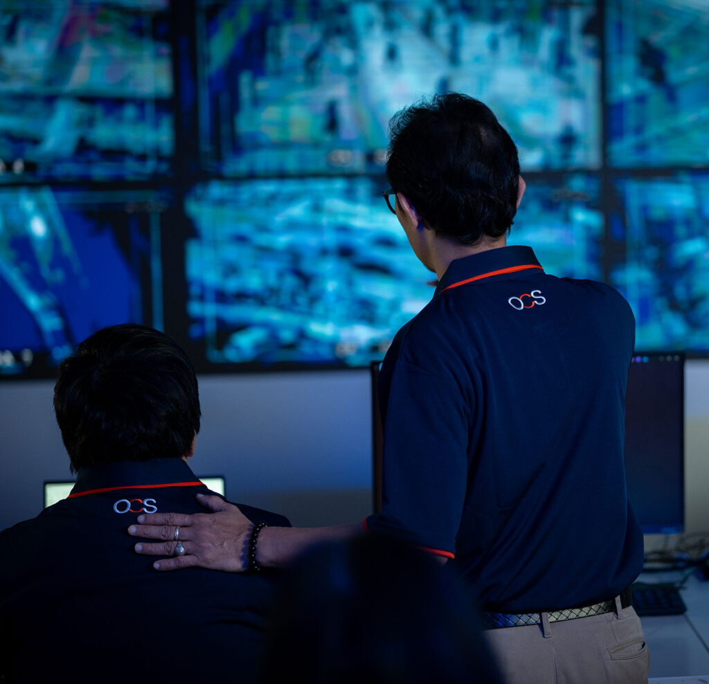 Two people in dark uniforms watch blue-tinted screens filled with data. One stands with a hand on the seated persons shoulder, both seen from behind. The atmosphere is focused and professional.