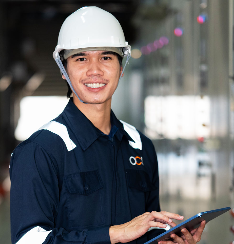 A smiling worker in a white hard hat and dark uniform holds a tablet in an industrial setting.