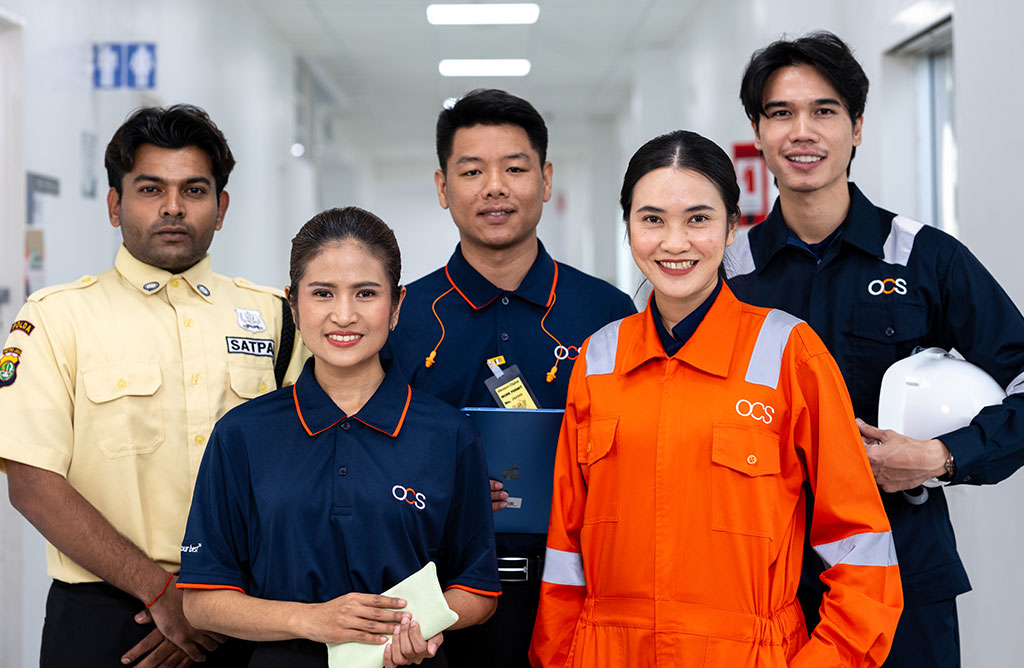 Five professionals in uniforms stand in a hallway, smiling at the camera. They represent different roles, including security and engineering, and are diverse in both gender and ethnicity.