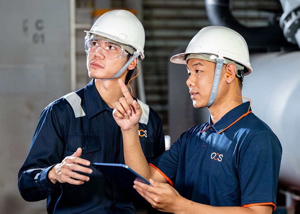 Two workers in helmets and safety glasses stand indoors; one points while the other holds a tablet, both looking in the same direction, appearing focused and engaged in discussion.