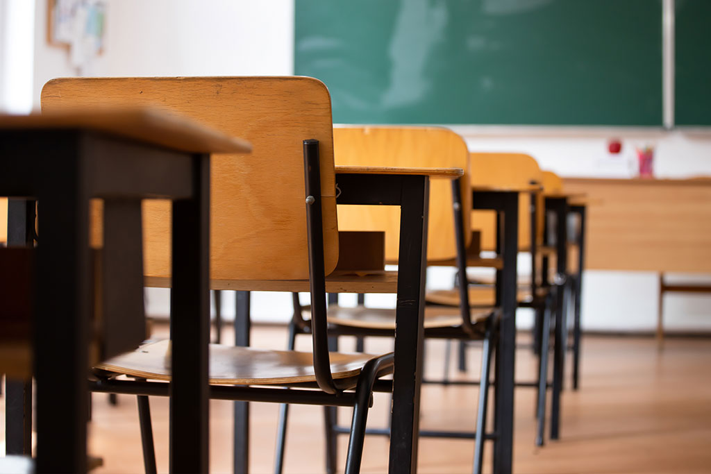 Rows of empty wooden desks and chairs in a classroom, with a green chalkboard and a teachers desk visible at the front. The room is well-lit with natural light.