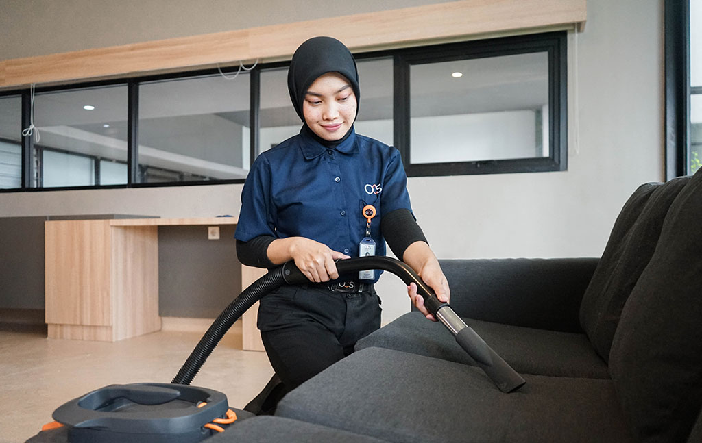 A woman wearing a black hijab and navy uniform vacuums a dark gray sofa in a modern, clean room with large windows and light wood furniture.
