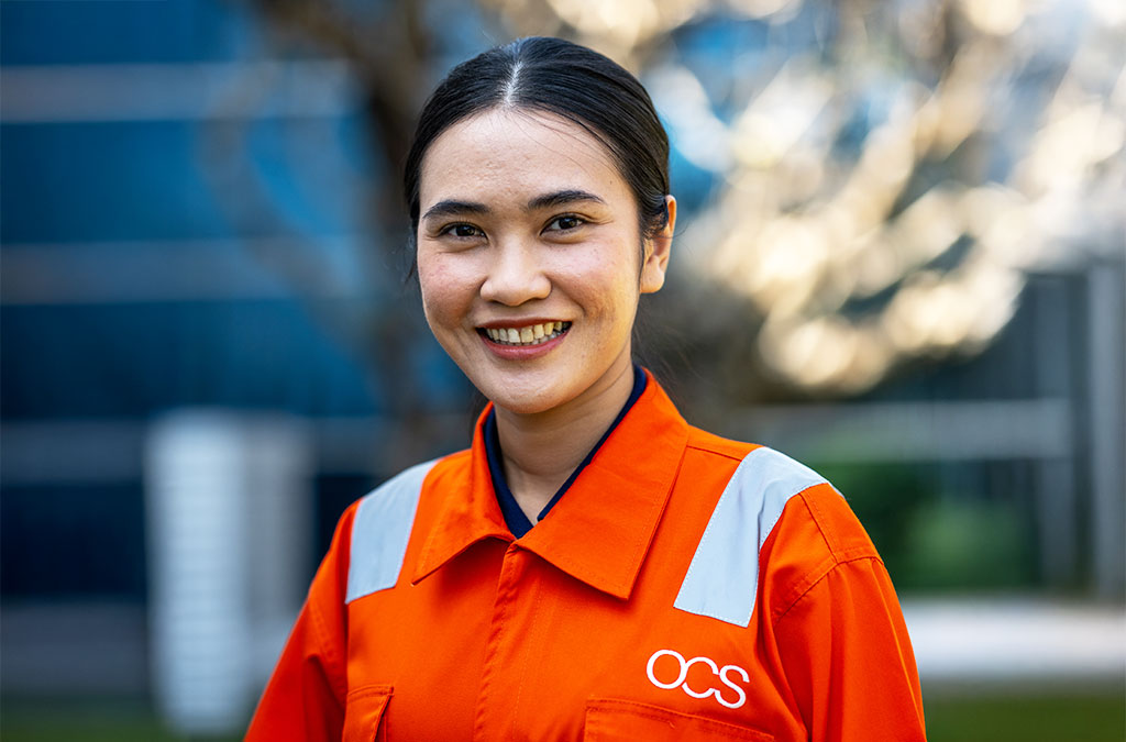 A woman in an orange uniform with reflective stripes and an OCS logo smiles outdoors, with a blurred background of trees and a building.