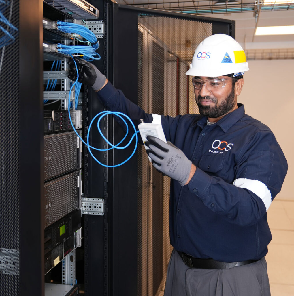 A technician in safety gear works on network cables in a server room, holding a device and inspecting connections.