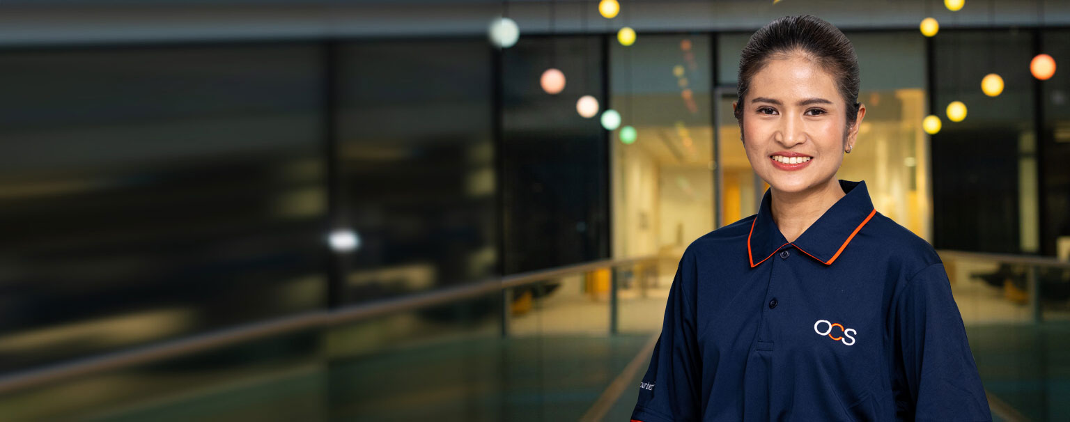 A woman in a navy OCS-branded polo shirt smiles, standing indoors with blurred colored lights and a modern glass background.